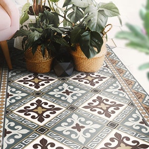 Op de afbeelding: Een vloerkleed met een patroon met een geometrisch ontwerp in blauw, bruin en wit. Het kleed heeft een rand met een bruin en gouden patroon. Het kleed ligt op een betegelde vloer met planten in potten op de achtergrond.