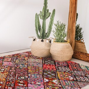 Peut inclure: Un tapis patchwork coloré aux tons rouges, roses, jaunes et bleus. Le tapis est posé sur un sol blanc devant un miroir en bois et deux plantes en pot.