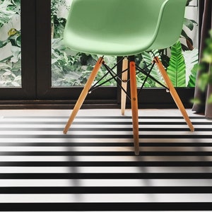 Puede incluir: Una silla verde con patas de madera se encuentra sobre una alfombra de rayas blancas y negras. La silla está situada frente a una ventana con plantas verdes visibles en el exterior.