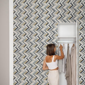 Può includere: Una persona con un top bianco e pantaloni beige è in piedi davanti a un armadio aperto. La parete dietro di lei presenta un motivo a zig-zag nei toni del grigio, giallo e nero. All'interno dell'armadio, i vestiti sono appesi a grucce di legno.