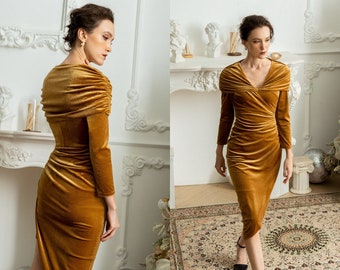 Puede incluir: Un vestido largo de terciopelo dorado con cuello de pico y silueta ajustada. El vestido tiene un ligero dobladillo asimétrico y se muestra en una modelo en un estudio.