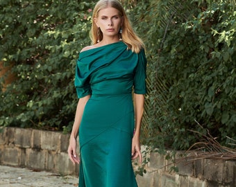 May include: A woman wearing a long, emerald green dress with a draped neckline and short sleeves. The dress has a fitted bodice and a flowing skirt. The woman is standing on a brick pathway with greenery in the background. The text "THE LINEN ATELIER" is visible at the bottom of the image.