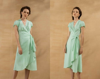 May include: A light green linen wrap dress with short sleeves and a tie at the waist. The dress is knee-length and has a flowing skirt. The model is wearing the dress with sandals. The text "THE LINEN ATELIER" is printed at the bottom of the image.