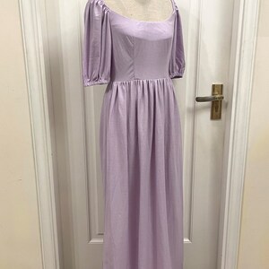 Puede incluir: Un vestido de lino lavanda con escote cuadrado y mangas abullonadas. El vestido tiene un corpi&ntilde;o ajustado y una falda fluida. El vestido se muestra en un maniqu&iacute; contra una puerta blanca.