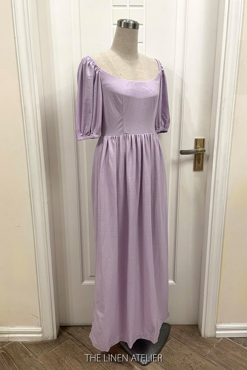 Puede incluir: Un vestido de lino lavanda con escote cuadrado y mangas abullonadas. El vestido tiene un corpi&ntilde;o ajustado y una falda fluida. El vestido se muestra en un maniqu&iacute; contra una puerta blanca.