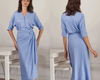 May include: A light blue linen wrap dress with a button-up front and a tie waist. The dress is knee-length and has a relaxed fit. The model is wearing the dress with a natural makeup look and her hair is styled in loose waves. The text "THE LINEN ATELIER" is visible at the bottom of the image.
