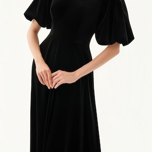 以下が含まれることがあります： オフショルダーネックラインと短いパフスリーブが特徴の黒いベルベットドレス。 ドレスはフィットしたボディスと流れるようなスカートです。 画像の下部にはThe Linen Atelierと印刷されています。