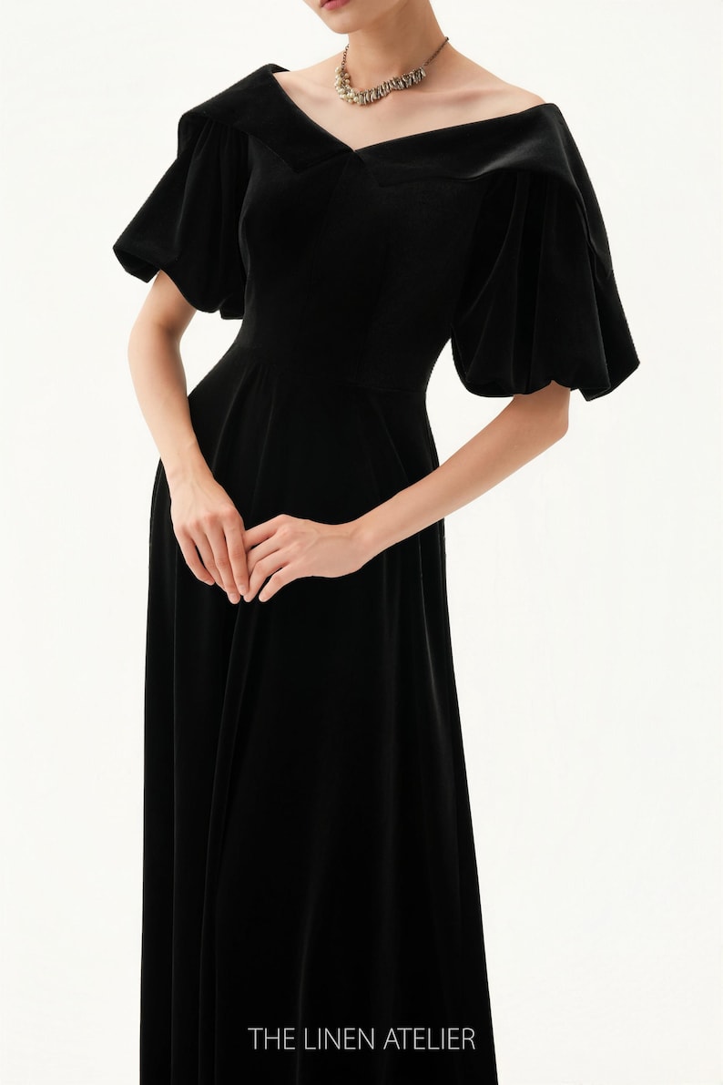 以下が含まれることがあります： オフショルダーネックラインと短いパフスリーブが特徴の黒いベルベットドレス。 ドレスはフィットしたボディスと流れるようなスカートです。 画像の下部にはThe Linen Atelierと印刷されています。