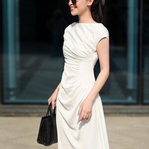 May include: A woman wearing a white dress and sunglasses is holding a black handbag. The dress has a gathered detail at the waist and a slightly asymmetrical hemline. The text "THE LINEN ATELIER" is visible at the bottom of the image.