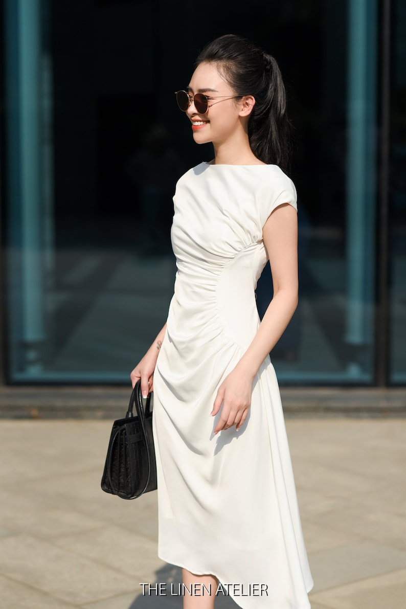 May include: A woman wearing a white dress and sunglasses is holding a black handbag. The dress has a gathered detail at the waist and a slightly asymmetrical hemline. The text "THE LINEN ATELIER" is visible at the bottom of the image.