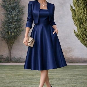 May include: A navy blue dress and jacket ensemble. The dress features a square neckline and a flared skirt. The cropped jacket has three-quarter length sleeves. A gold clutch and navy heels complete the look. The outfit is suitable for formal events.