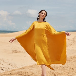 Puede incluir: Una mujer con un vestido amarillo mostaza de manga larga y dise&ntilde;o de capa, de pie en una playa de arena. El vestido llega a la rodilla. El texto "THE LINEN ATELIER" es visible en la parte inferior de la imagen, con arena y cielo de fondo.