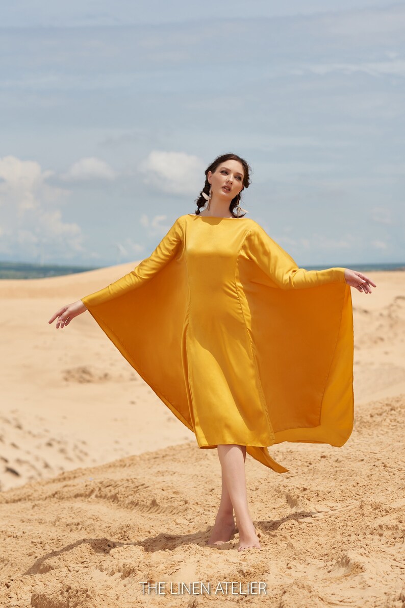 Puede incluir: Una mujer con un vestido amarillo mostaza de manga larga y dise&ntilde;o de capa, de pie en una playa de arena. El vestido llega a la rodilla. El texto "THE LINEN ATELIER" es visible en la parte inferior de la imagen, con arena y cielo de fondo.