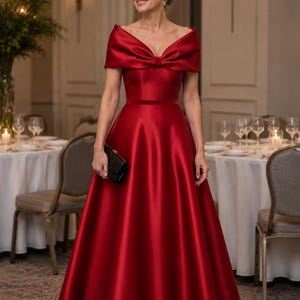Può includere: Un abito formale in raso rosso con scollatura a spalle scoperte e dettaglio a fiocco. L'abito ha un corpetto aderente e una gonna ampia e fluida. Viene tenuta una pochette nera. Il testo "THE LINEN ATELIER" è in basso nell'immagine.