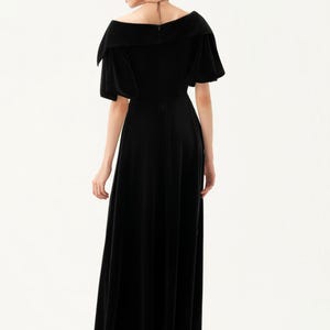 以下が含まれることがあります： オフショルダーネックラインと短いパフスリーブが特徴の黒いベルベットのマキシドレス。ドレスはフィットしたボディスと流れるようなスカートです。画像の下部にはThe Linen Atelierと印刷されています。