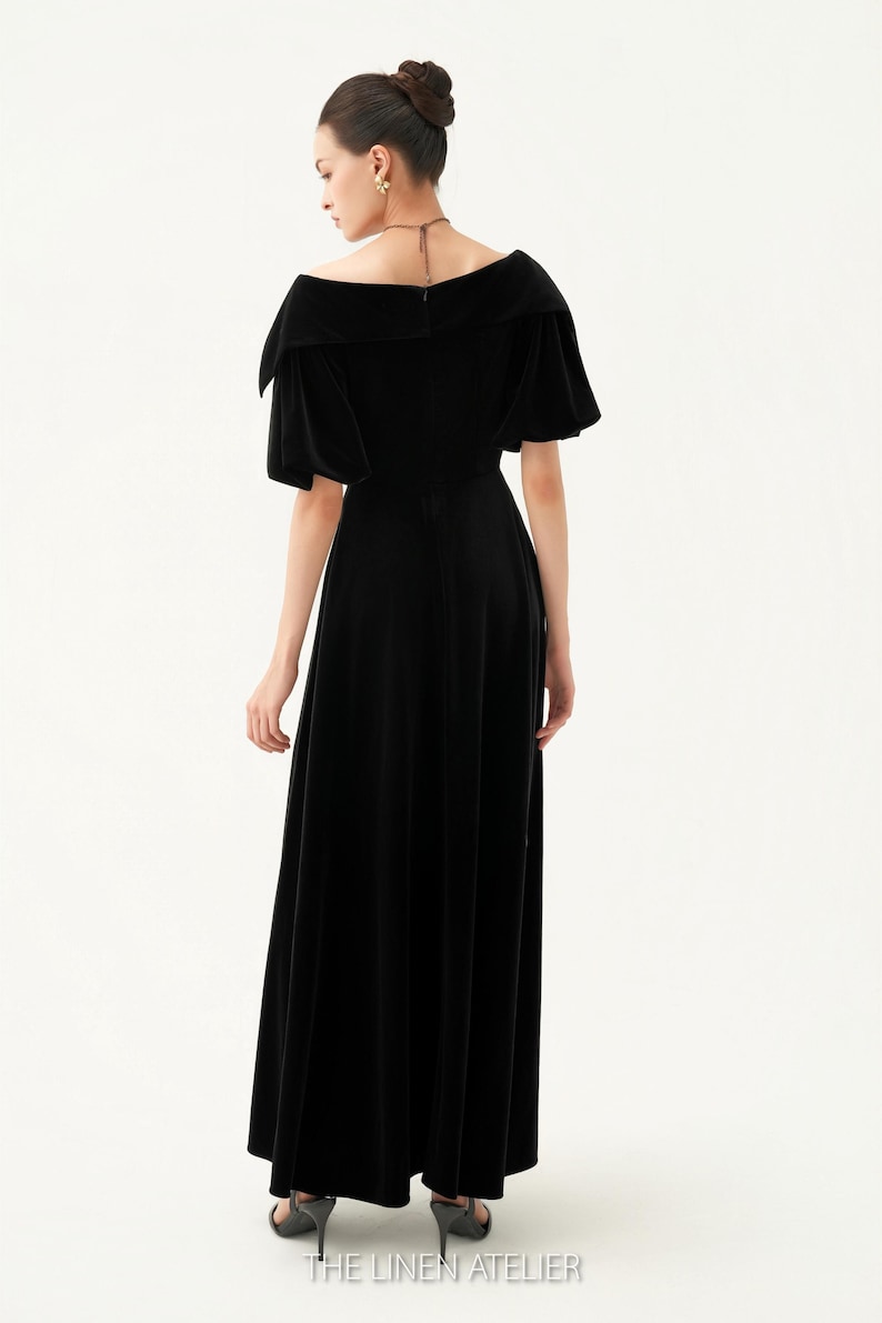 以下が含まれることがあります： オフショルダーネックラインと短いパフスリーブが特徴の黒いベルベットのマキシドレス。ドレスはフィットしたボディスと流れるようなスカートです。画像の下部にはThe Linen Atelierと印刷されています。
