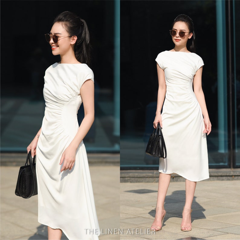 May include: A white midi dress with a gathered waist and short sleeves. The dress is made of a lightweight fabric and has a relaxed fit. The model is wearing the dress with a pair of black sunglasses and a black handbag.