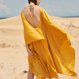 Puede incluir: Vestido amarillo mostaza fluido, con espalda baja y mangas largas, usado al aire libre en una playa de arena. El tejido ondea con el viento. El texto "THE LINEN ATELIER" es visible en la parte inferior de la imagen.