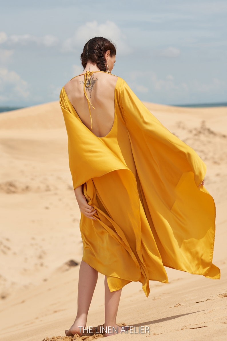Puede incluir: Vestido amarillo mostaza fluido, con espalda baja y mangas largas, usado al aire libre en una playa de arena. El tejido ondea con el viento. El texto "THE LINEN ATELIER" es visible en la parte inferior de la imagen.