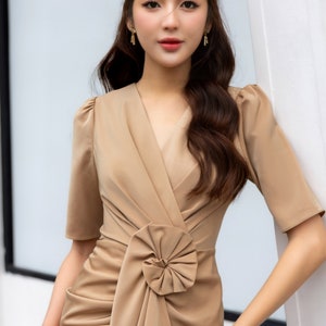 May include: A woman wearing a brown, short-sleeved dress with a large bow detail at the waist. The dress is fitted and has a V-neckline. The woman is standing in front of a white wall. The text "THE LINEN ATELIER" is visible at the bottom of the image.