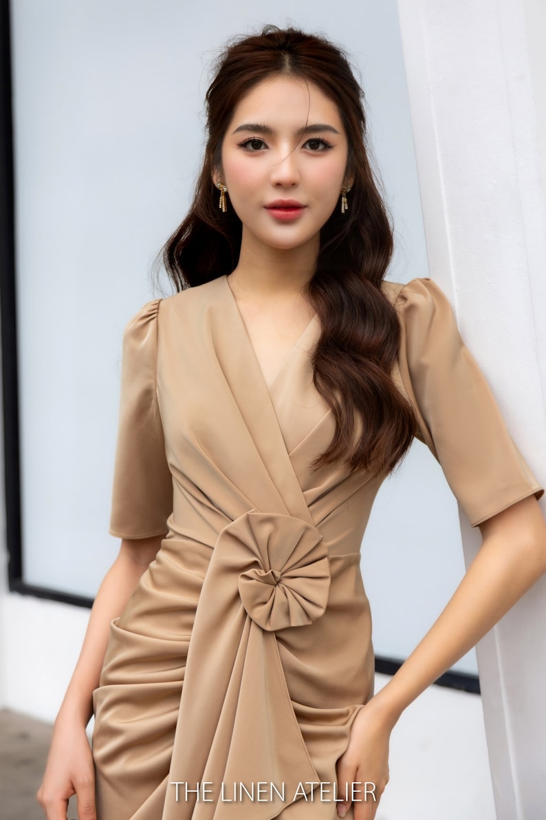 May include: A woman wearing a brown, short-sleeved dress with a large bow detail at the waist. The dress is fitted and has a V-neckline. The woman is standing in front of a white wall. The text "THE LINEN ATELIER" is visible at the bottom of the image.