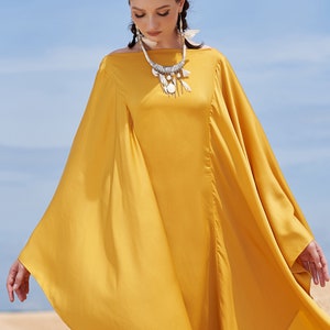 Puede incluir: Vestido fluido amarillo mostaza con mangas anchas y cuello barco. El vestido se complementa con un collar plateado con detalles blancos. El fondo muestra una playa de arena y un cielo azul. El texto "THE LINEN ATELIER" es visible en la parte inferior.