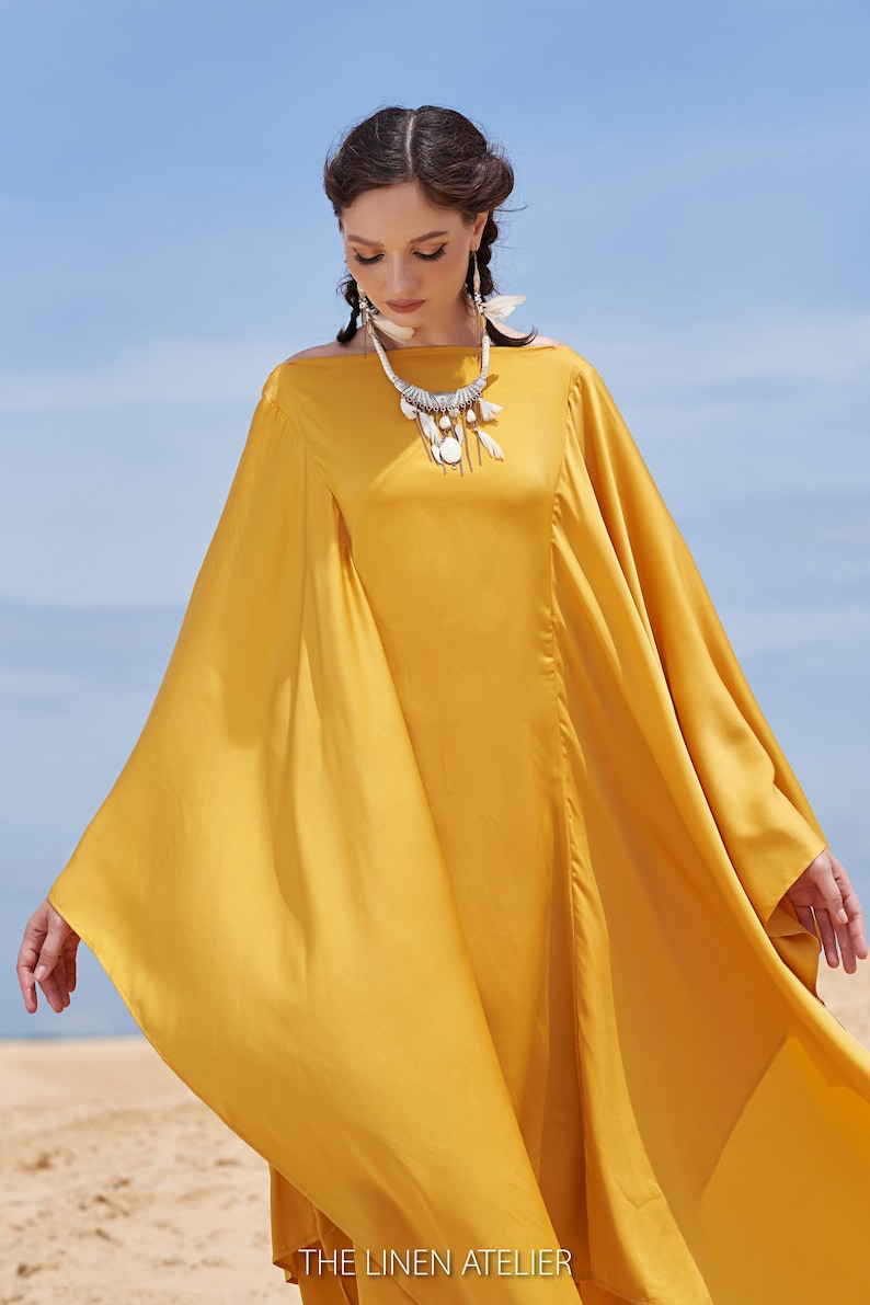 Puede incluir: Vestido fluido amarillo mostaza con mangas anchas y cuello barco. El vestido se complementa con un collar plateado con detalles blancos. El fondo muestra una playa de arena y un cielo azul. El texto "THE LINEN ATELIER" es visible en la parte inferior.