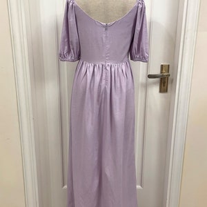 Puede incluir: Un vestido de lino lavanda con escote cuadrado y mangas abullonadas. El vestido tiene una falda fruncida y una cremallera en la espalda. El vestido est&aacute; en un maniqu&iacute; delante de una puerta blanca.