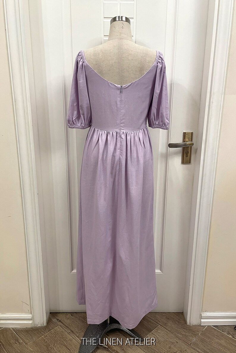 Puede incluir: Un vestido de lino lavanda con escote cuadrado y mangas abullonadas. El vestido tiene una falda fruncida y una cremallera en la espalda. El vestido est&aacute; en un maniqu&iacute; delante de una puerta blanca.