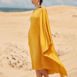 Puede incluir: Un vestido amarillo mostaza con mangas fluidas es usado por una persona de pie sobre una duna de arena. El vestido tiene un dise&ntilde;o simple y elegante. The Linen Atelier est&aacute; impreso en la parte inferior de la imagen.