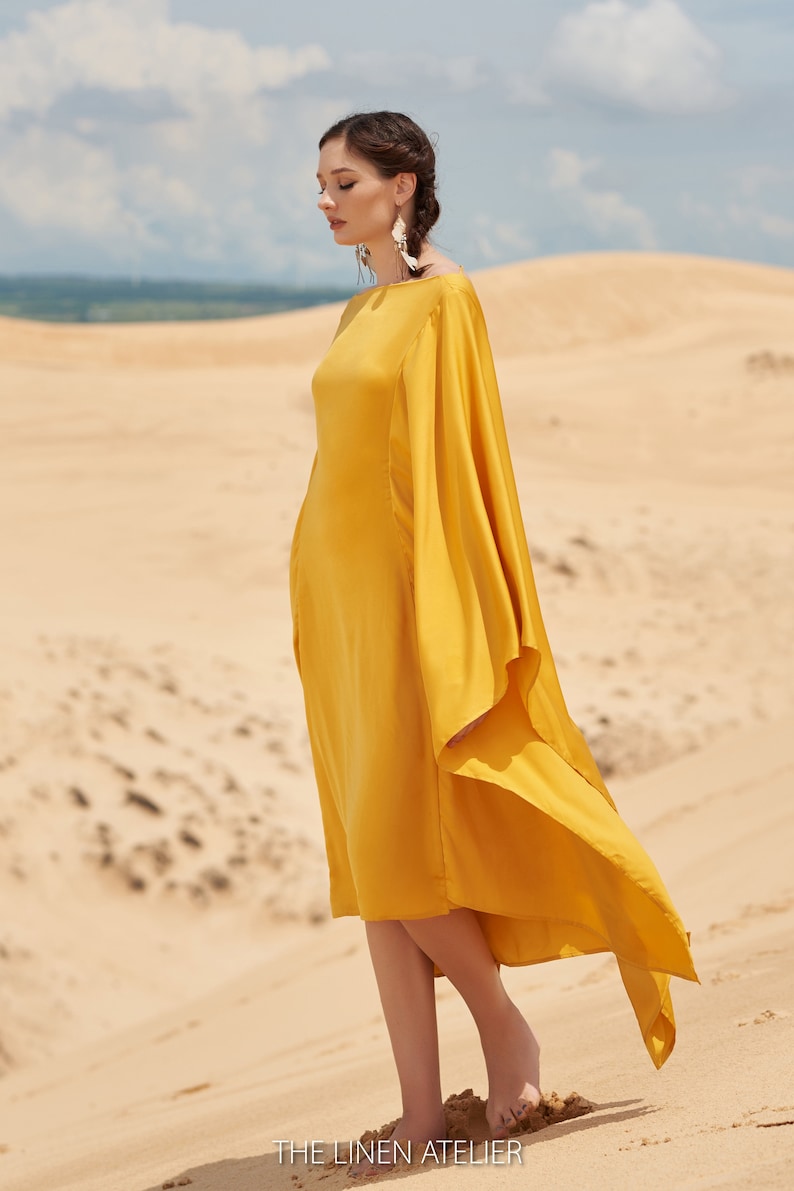 Puede incluir: Un vestido amarillo mostaza con mangas fluidas es usado por una persona de pie sobre una duna de arena. El vestido tiene un dise&ntilde;o simple y elegante. The Linen Atelier est&aacute; impreso en la parte inferior de la imagen.