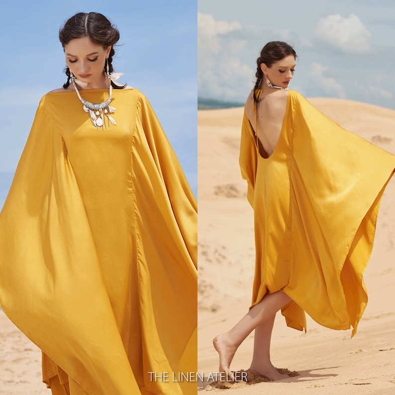 Puede incluir: Vestido amarillo mostaza fluido con mangas anchas y espalda baja, usado en una playa de arena. El logotipo de The Linen Atelier es visible en la parte inferior de la imagen. La modelo lleva un collar. El vestido es de tela suave.