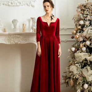 May include: A woman wearing a long, red velvet dress with a V-neckline and three-quarter sleeves. She is standing in front of a fireplace and a Christmas tree. The text "THE LINEN ATELIER" is at the bottom of the image.