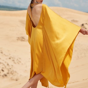 Puede incluir: Vestido amarillo mostaza fluido con espalda baja y mangas anchas. La modelo est&aacute; de pie sobre una superficie arenosa. El vestido es de THE LINEN ATELIER.