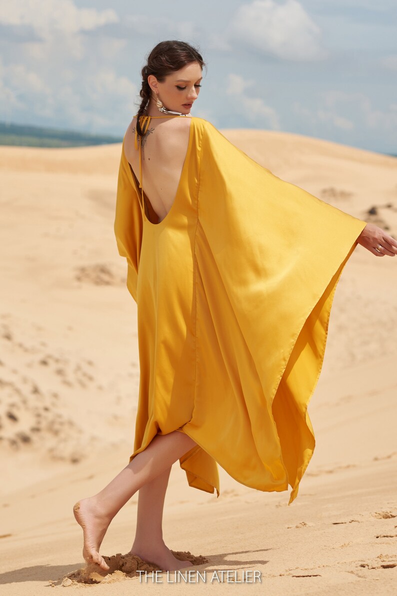 Puede incluir: Vestido amarillo mostaza fluido con espalda baja y mangas anchas. La modelo est&aacute; de pie sobre una superficie arenosa. El vestido es de THE LINEN ATELIER.