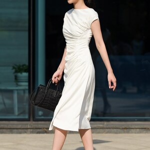 May include: A woman wearing a white dress and clear heeled sandals walks in front of a glass building. She is carrying a black handbag with a crocodile pattern. The text "THE LINEN ATELIER" is visible at the bottom of the image.