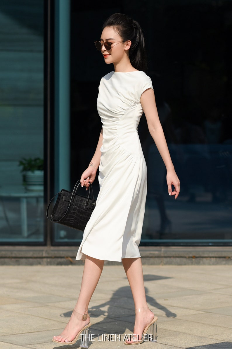 May include: A woman wearing a white dress and clear heeled sandals walks in front of a glass building. She is carrying a black handbag with a crocodile pattern. The text "THE LINEN ATELIER" is visible at the bottom of the image.