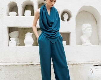 Könnte beinhalten: Ein blaugrüner Jumpsuit mit einem drapierten Schalkragen und weiten Beinen. Das Model steht vor einer weißen Wand mit dekorativen Nischen.