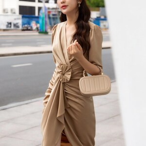 May include: A woman wearing a beige satin dress with a large bow detail at the waist. She is holding a small beige handbag with a gold clasp. The dress has a V-neckline and three-quarter sleeves. The woman is standing in front of a white wall. The text "THE LINEN ATELIER" is visible at the bottom of the image.