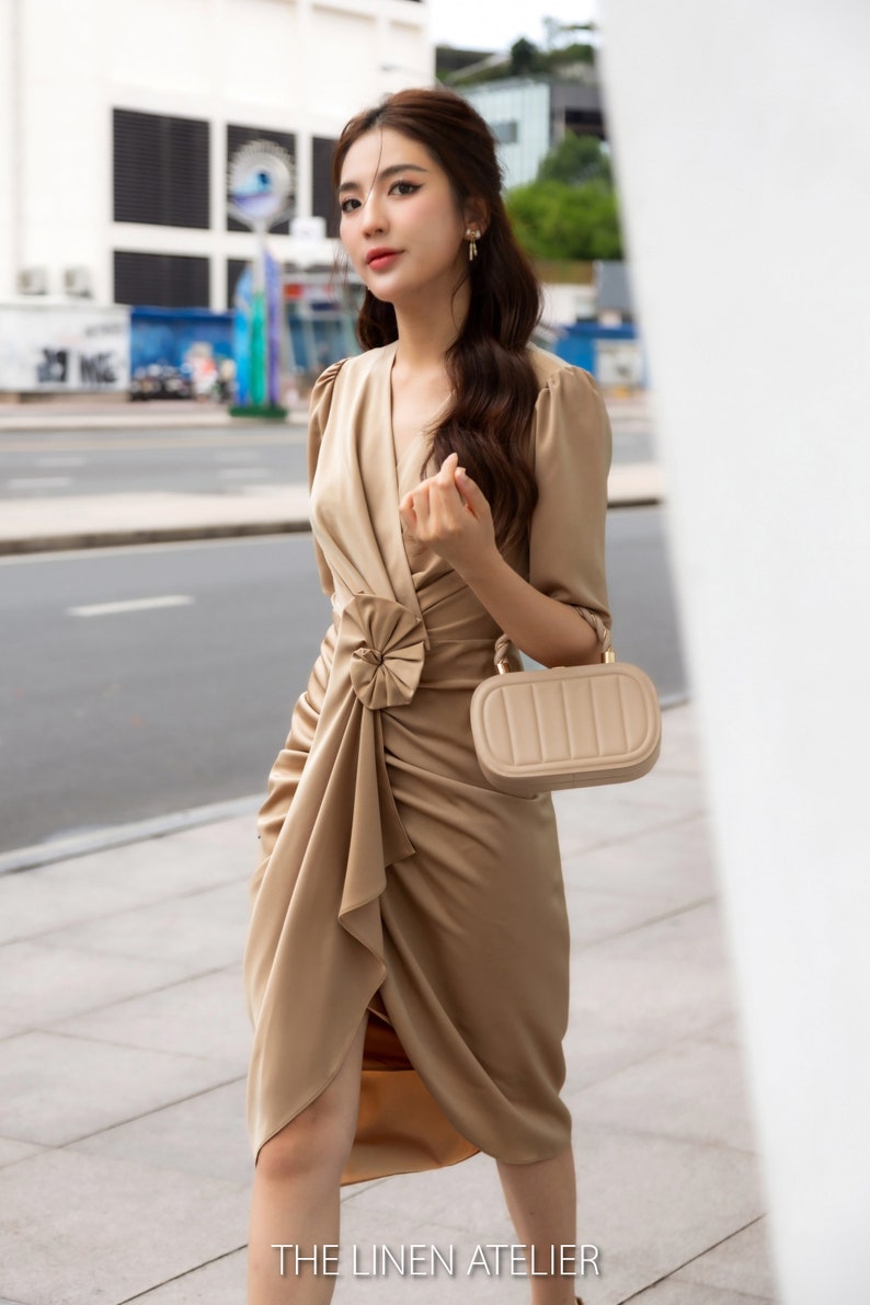 May include: A woman wearing a beige satin dress with a large bow detail at the waist. She is holding a small beige handbag with a gold clasp. The dress has a V-neckline and three-quarter sleeves. The woman is standing in front of a white wall. The text "THE LINEN ATELIER" is visible at the bottom of the image.