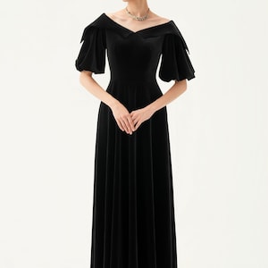 以下が含まれることがあります： オフショルダーの広いネックラインと短いパフスリーブが特徴の、黒のベルベットのロングドレス。 ドレスはフィットしたボディスと流れるようなスカートです。 下部には「THE LINEN ATELIER」の文字があります。