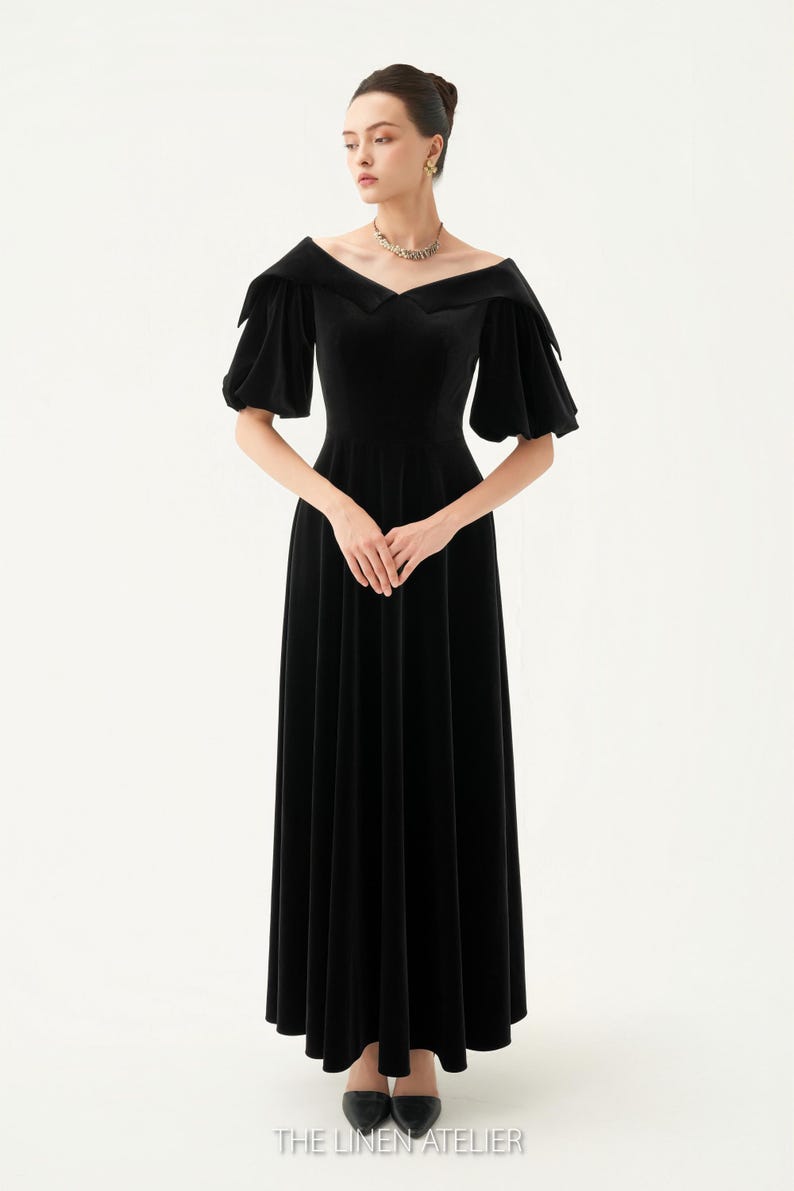 以下が含まれることがあります： オフショルダーの広いネックラインと短いパフスリーブが特徴の、黒のベルベットのロングドレス。 ドレスはフィットしたボディスと流れるようなスカートです。 下部には「THE LINEN ATELIER」の文字があります。