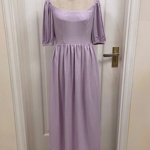 Puede incluir: Un vestido de lino lavanda con escote cuadrado y mangas abullonadas. El vestido tiene un corpi&ntilde;o ajustado y una falda fluida. El vestido est&aacute; en un maniqu&iacute; frente a una puerta blanca.
