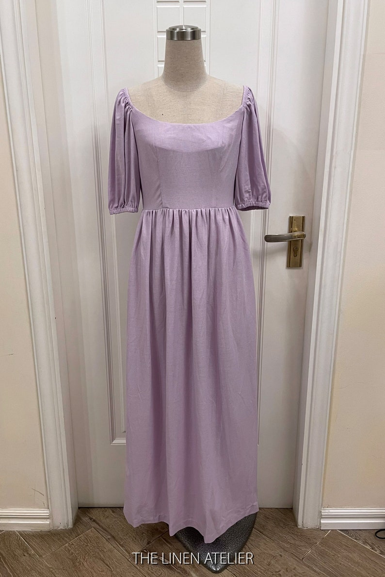 Puede incluir: Un vestido de lino lavanda con escote cuadrado y mangas abullonadas. El vestido tiene un corpi&ntilde;o ajustado y una falda fluida. El vestido est&aacute; en un maniqu&iacute; frente a una puerta blanca.