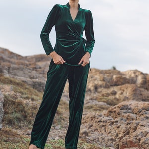 May include: A woman wearing a dark green velvet pantsuit with a wrap top and long sleeves. She is standing on a rocky hillside with green grass.