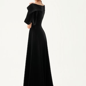 以下が含まれることがあります： 黒いベルベットのロングドレス。広いネックラインと短い袖が特徴です。ドレスはAラインスカートで、黒いヒールと合わせています。画像の下部には「THE LINEN ATELIER」と印刷されています。