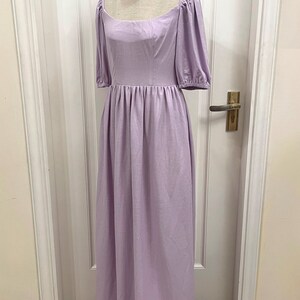 Puede incluir: Un vestido de lino lavanda con escote cuadrado y mangas abullonadas. El vestido tiene un corpi&ntilde;o ajustado y una falda fluida. El vestido est&aacute; en un maniqu&iacute; frente a una puerta blanca.