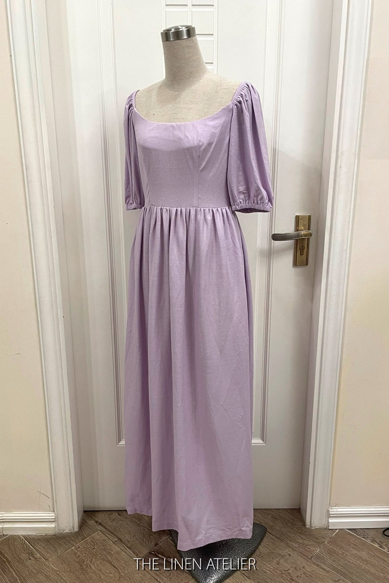 Puede incluir: Un vestido de lino lavanda con escote cuadrado y mangas abullonadas. El vestido tiene un corpi&ntilde;o ajustado y una falda fluida. El vestido est&aacute; en un maniqu&iacute; frente a una puerta blanca.
