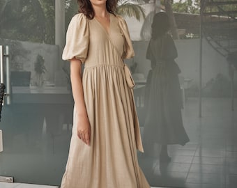 May include: A beige linen wrap dress with a V-neckline and puffed sleeves. The dress has a tie at the waist and falls to the mid-calf. The model is wearing the dress with nude heels. The text "THE LINEN ATELIER" is at the bottom of the image.