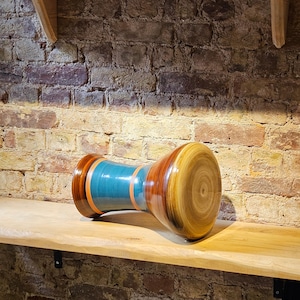 May include: A handcrafted wooden drum with a teal central section, bordered by orange and brown bands. The drum's ends are light brown with concentric circular patterns. The drum is displayed on a wooden shelf against a brick wall.